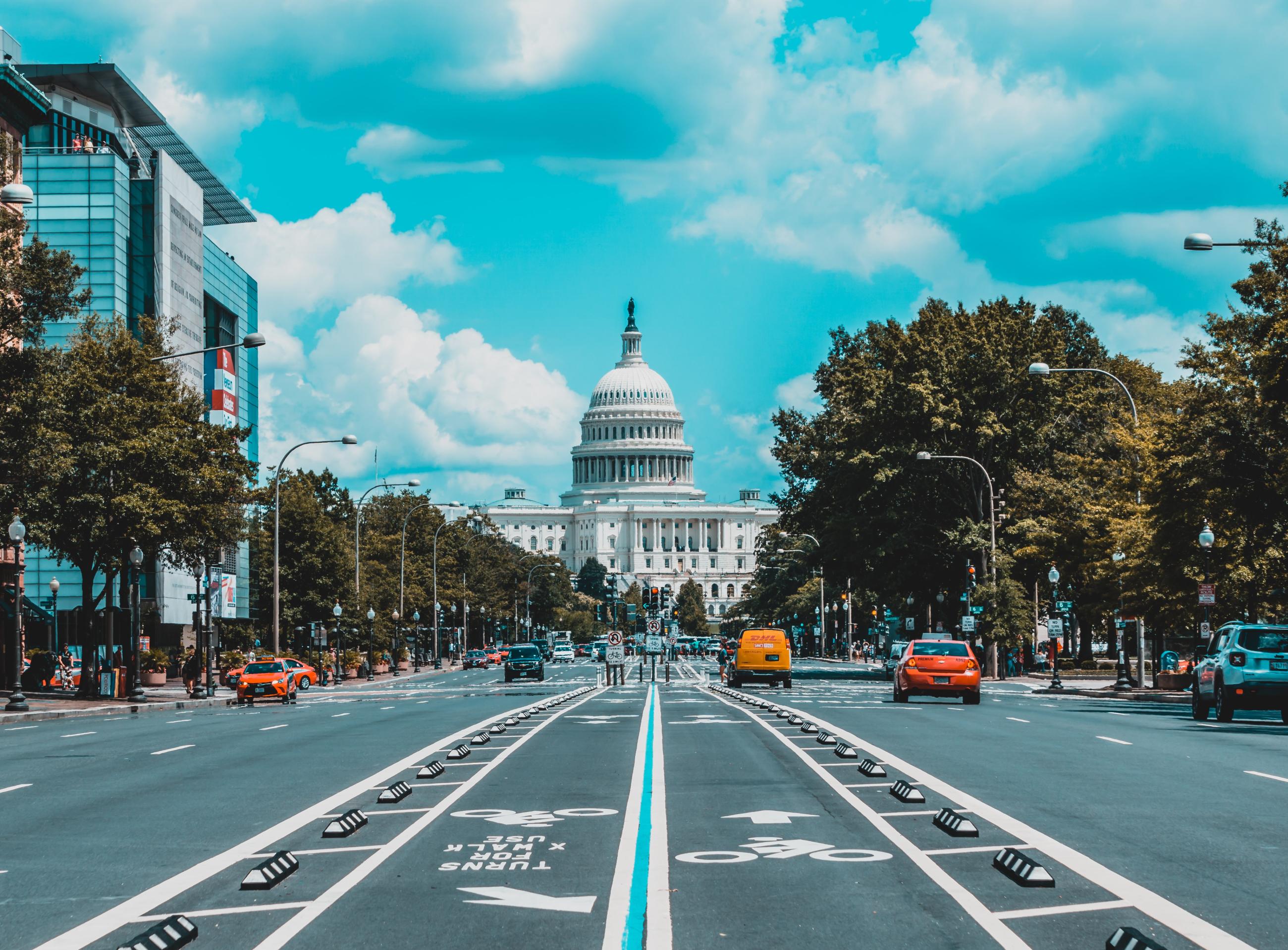 A view looking down Pennsylvania Avenue toward the U.S. Capitol building in Washington, D.C., on a sunny day with bright blue skies and scattered clouds. The wide boulevard features marked bike lanes and a center median, with cars and a yellow DHL delivery truck in traffic. Tree-lined sidewalks and commercial buildings flank both sides of the street.