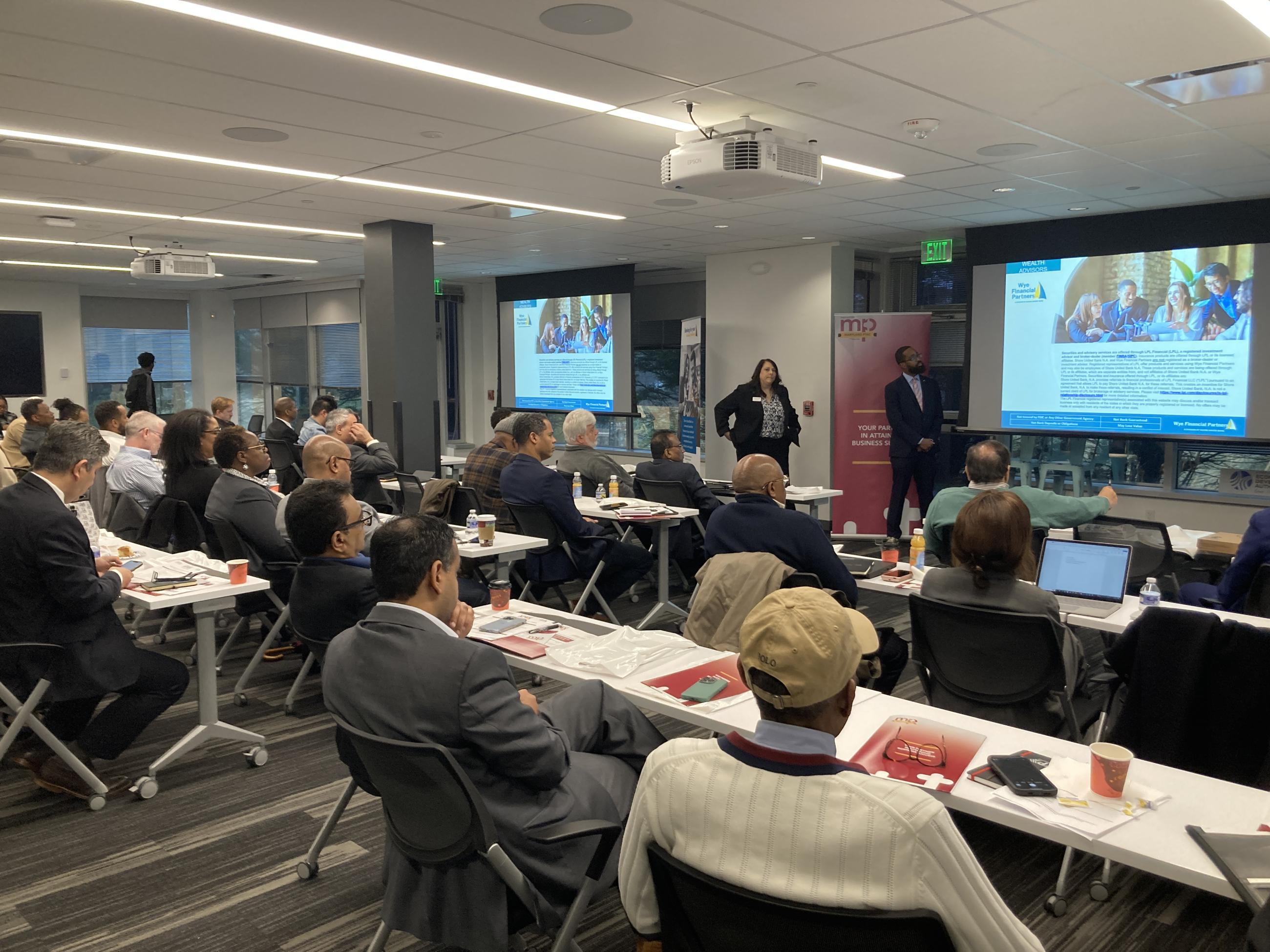 Attendees at a Maryland APEX Accelerator event listen to two presenters during a business development workshop, with projected slides and a Maryland APEX banner visible at the front of the room.