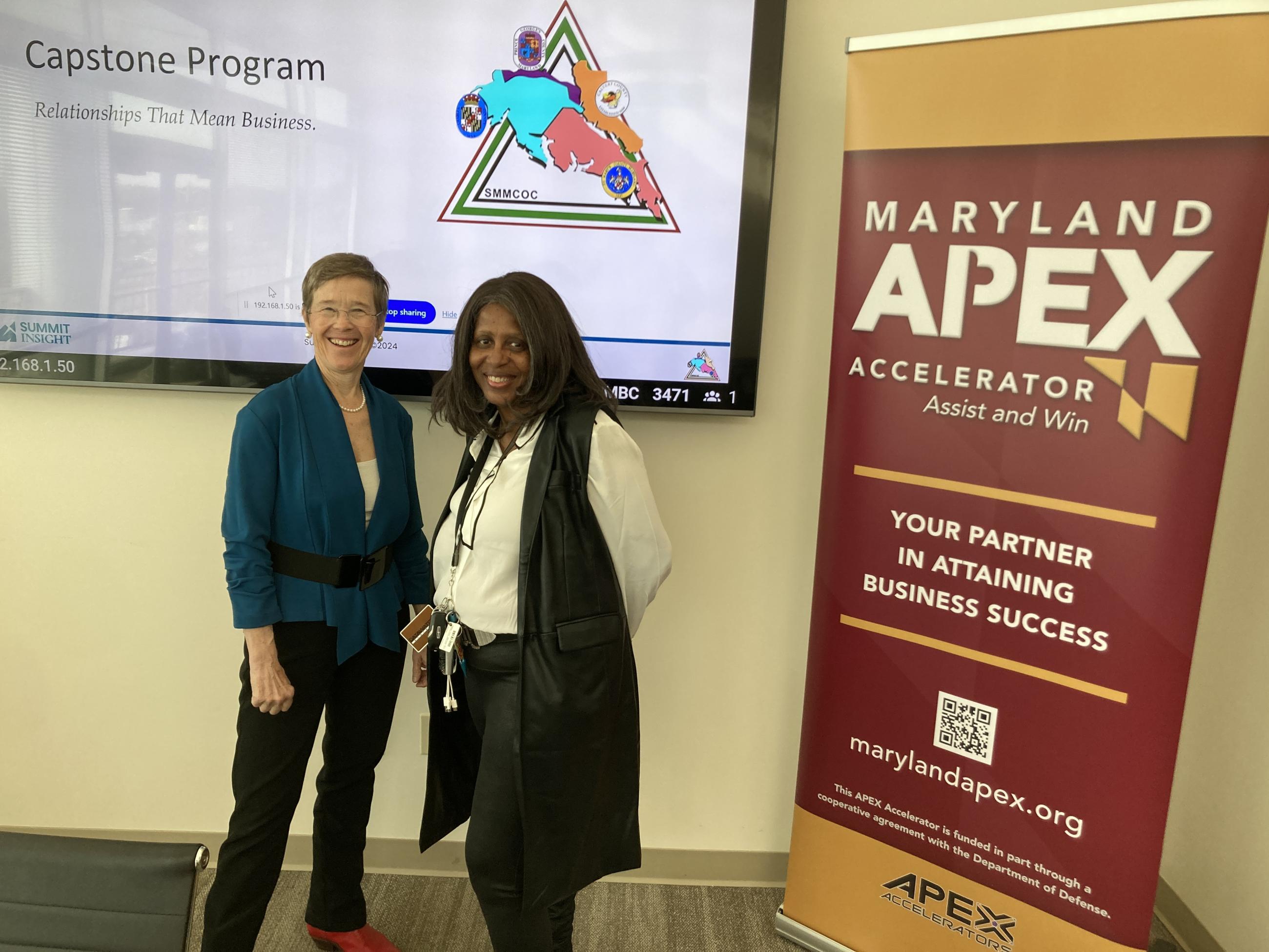 Two women smiling together at a Maryland APEX Accelerator Capstone Program event titled 'Relationships That Mean Business,' with a presentation screen displaying the SMMCOC logo and a Maryland APEX Accelerator 'Your Partner in Attaining Business Success' banner beside them