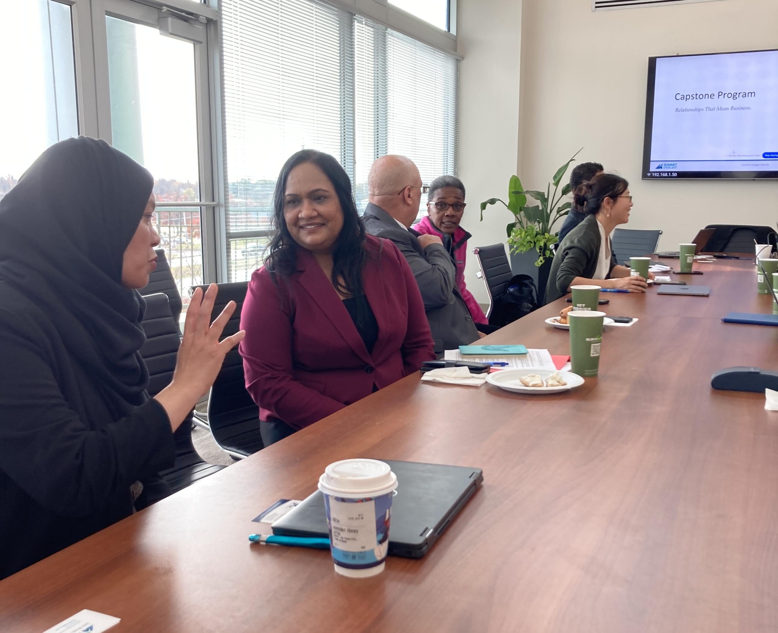 A small group of professionals seated around a large conference table during a Maryland APEX Accelerator Capstone Program session, with a 'Relationships That Mean Business' presentation visible on a screen in the background. Two women in the foreground are engaged in conversation.