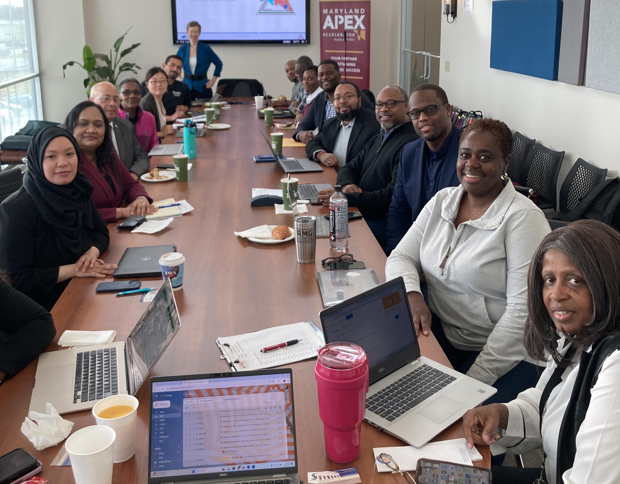 A large group of approximately 16 professionals seated around a long conference table during a Maryland APEX Accelerator Capstone Program meeting, with laptops, notes, and beverages on the table. A presenter stands at the head of the room in front of a screen displaying the Capstone Program branding, with a Maryland APEX Accelerator banner visible to the right