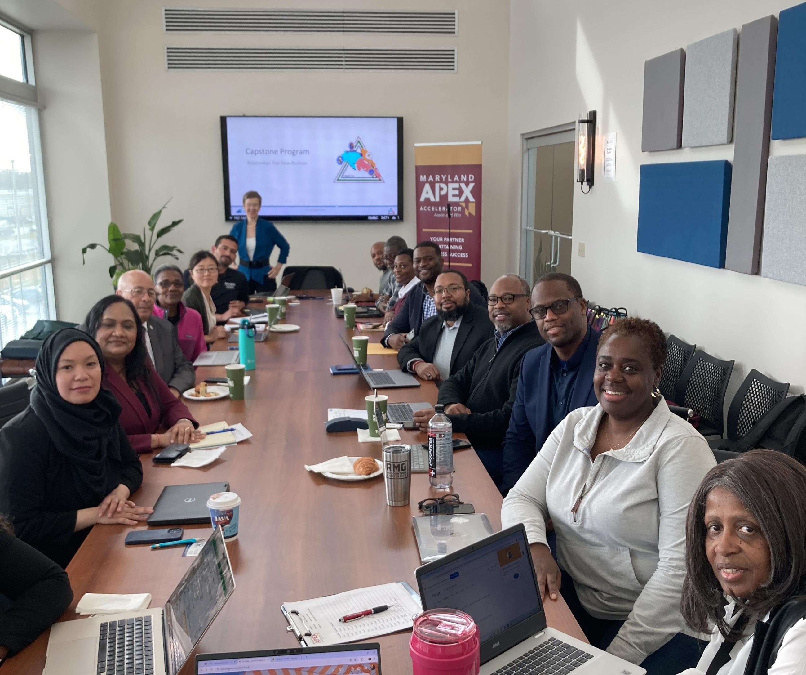 A diverse group of professionals seated around a large conference table with laptops and notebooks, attending a Capstone Program training session, with a Maryland APEX Accelerator banner visible in the background