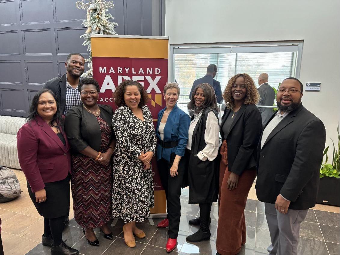 Eight professionals posing together in a building lobby at a Maryland APEX Accelerator holiday event, with a decorated Christmas tree and a Maryland APEX Accelerator pull-up banner visible in the background.