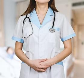 Healthcare professional in a white medical uniform with light blue collar and cuff trim, wearing a stethoscope around their neck, with hands clasped in front, standing in a hospital setting.